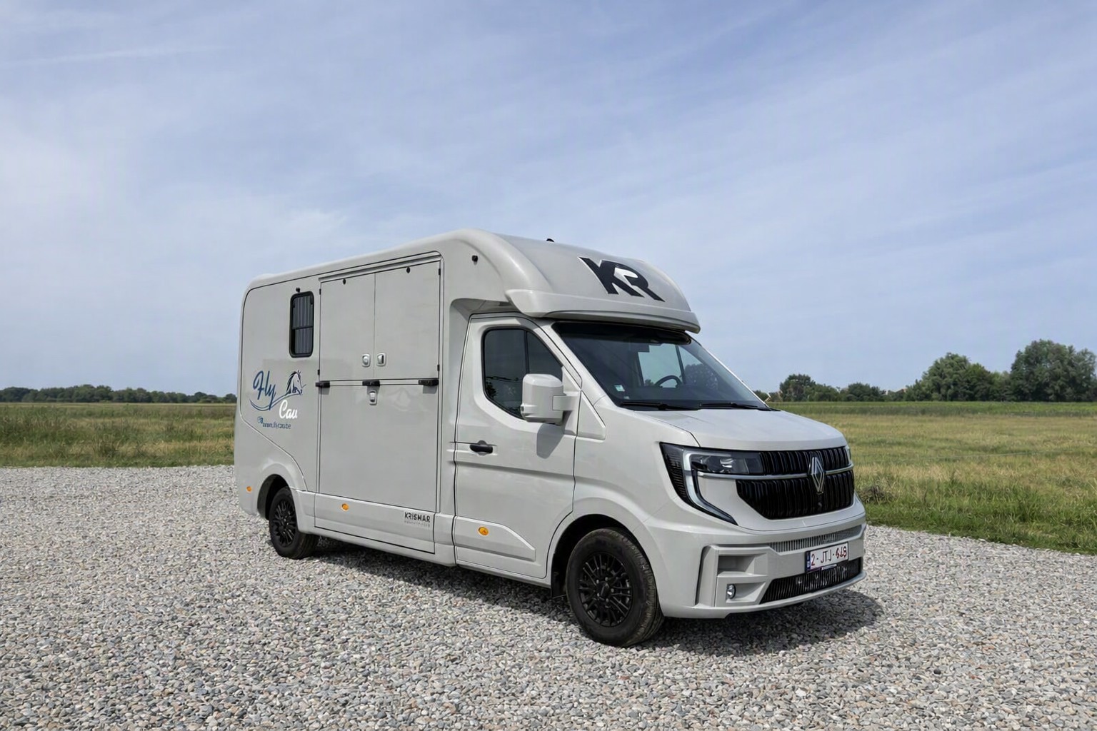 Witte Renault Master Krismar-paardencamionette met het FLY CAV-logo, geparkeerd op een grindterrein in een open landelijke omgeving. Op de wagen staan de teksten Fly Cav, www.flycav.be en Krismar Horsetrucks. De foto heeft een moderne en professionele uitstraling.